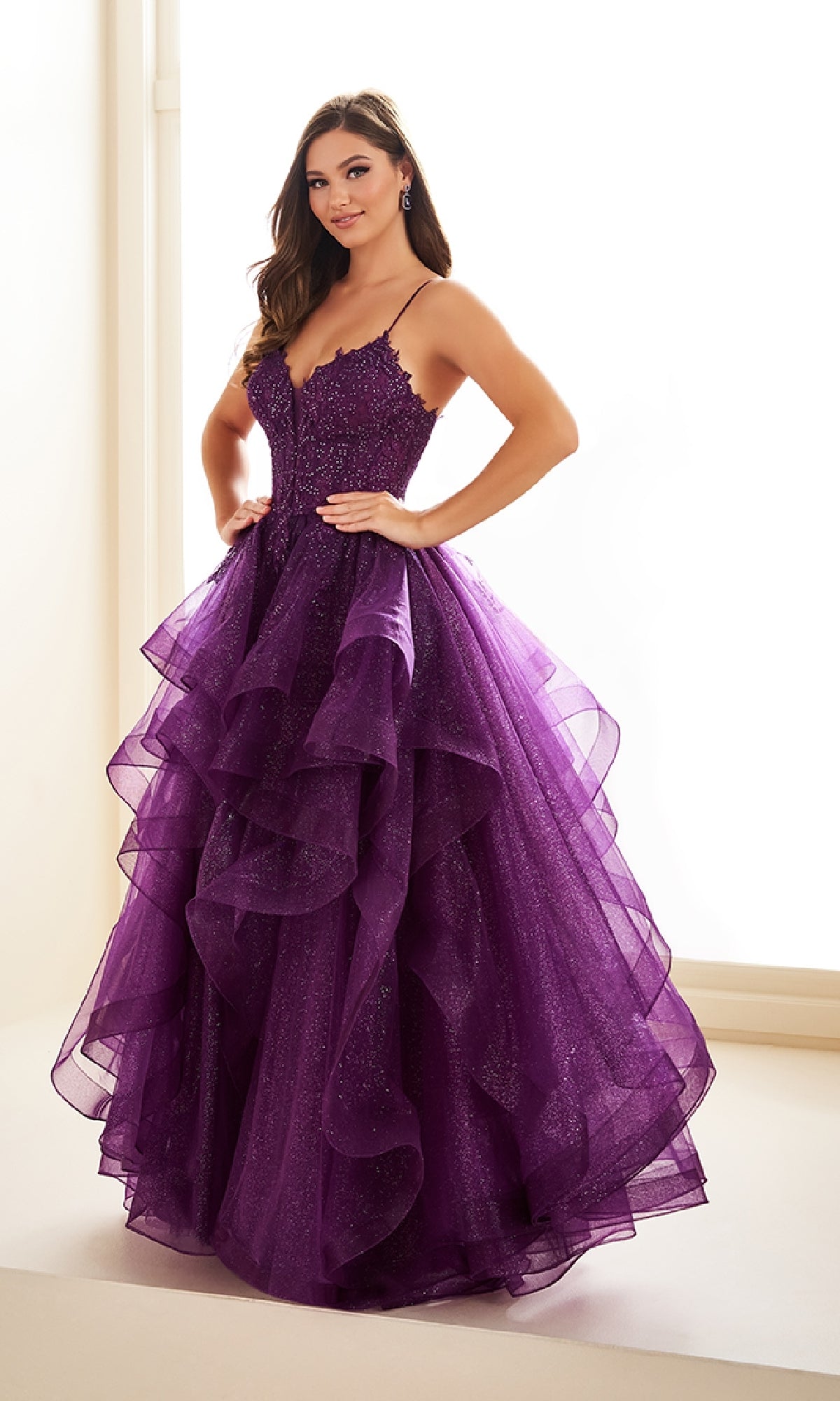 A woman stands indoors with hands on hips, wearing the Ellie Wilde EW36203 glitter tulle prom ball gown featuring a purple layered skirt and an embroidered corset bodice.
